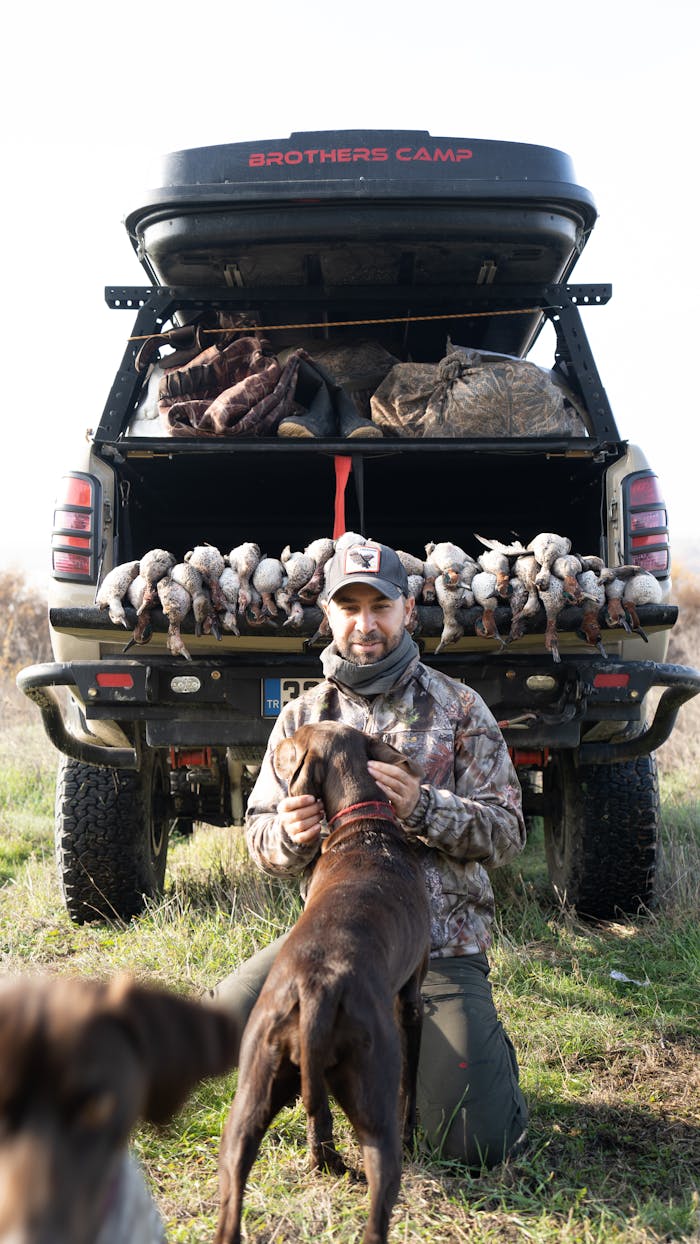 A hunter poses with his dog and harvested ducks at the back of a pickup truck.